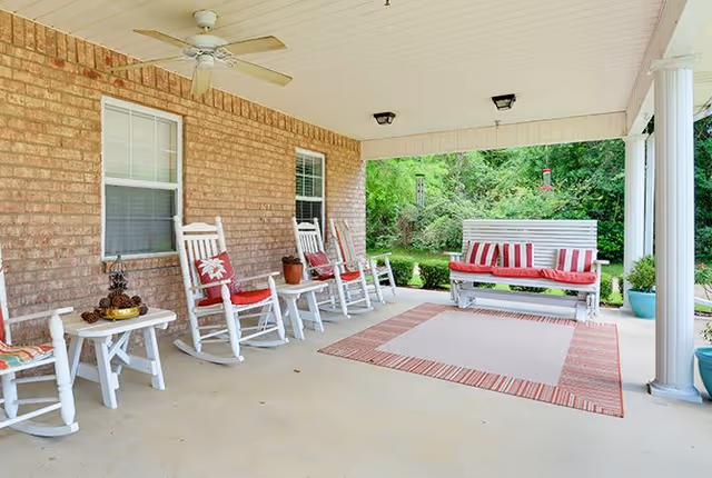 Covered outdoor porch area with white rocking chairs and a bench with red and white striped cushions. The porch has a ceiling fan, two windows on a brick wall, a small table with decorative items, and a large outdoor rug. Greenery and trees are visible beyond the porch.