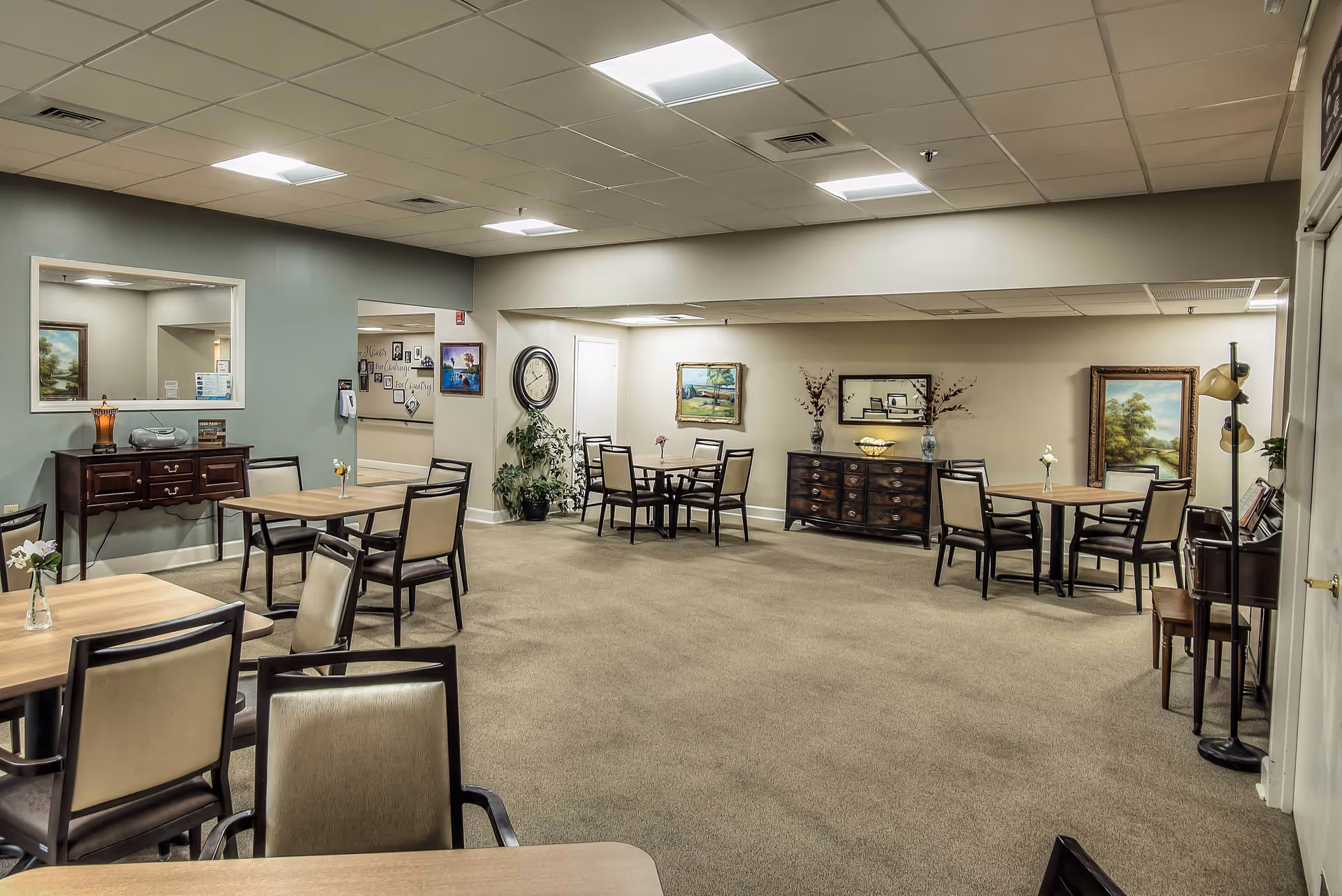 A spacious common area with multiple tables and chairs arranged for seating. The room has beige carpeting, light-colored walls, and a drop ceiling with fluorescent lighting. There are decorative elements including framed paintings, a large wall clock, plants, and a wooden sideboard with vases and a bowl. A large mirror is mounted on one wall, and a floor lamp stands in the corner.