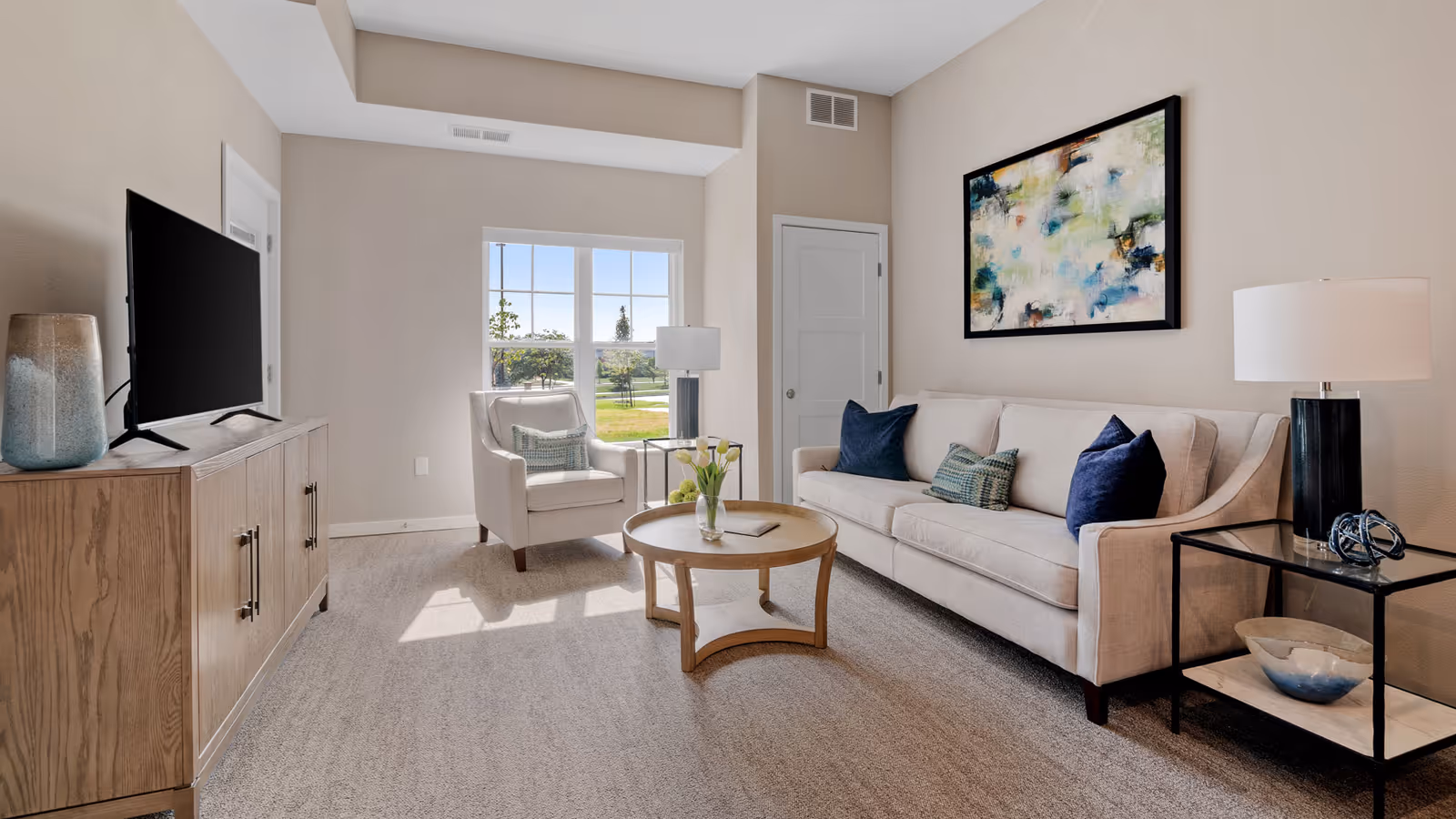 Bright furnished living room with a sofa, armchair, coffee table, TV on a wooden console and a window showing greenery outside.