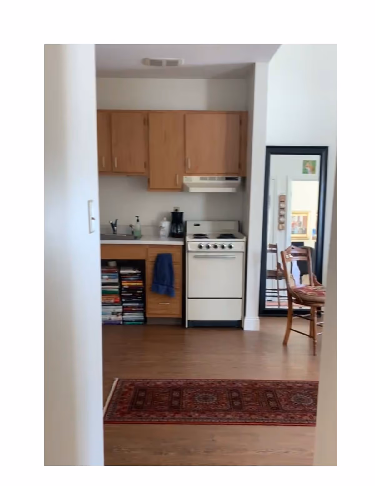 Small kitchenette with wooden cabinets, a stove and sink, bookshelves under the counter and a chair with a tall mirror to the right.