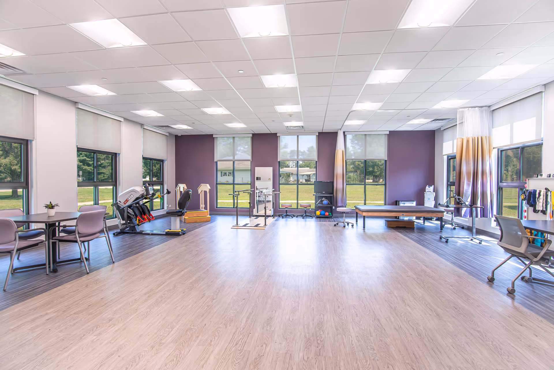 A spacious, well-lit rehabilitation or physical therapy room with large windows showing green outdoor scenery. The room features exercise equipment including a recumbent bike, parallel bars, therapy balls, and a treatment table. There are also chairs and tables on the left side and a water cooler on the right side.