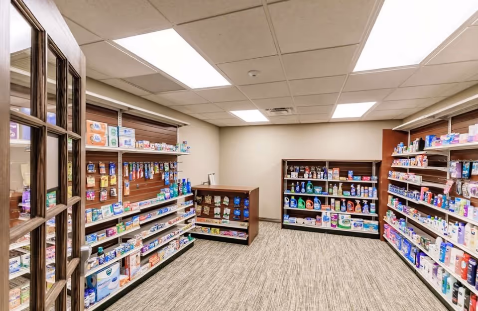 Interior view of a small retail or convenience store area within a facility, featuring shelves stocked with various personal care products, household items, and over-the-counter medications. The room has a beige carpet, white ceiling tiles with fluorescent lighting, and a wooden framed glass door partially open on the left side.