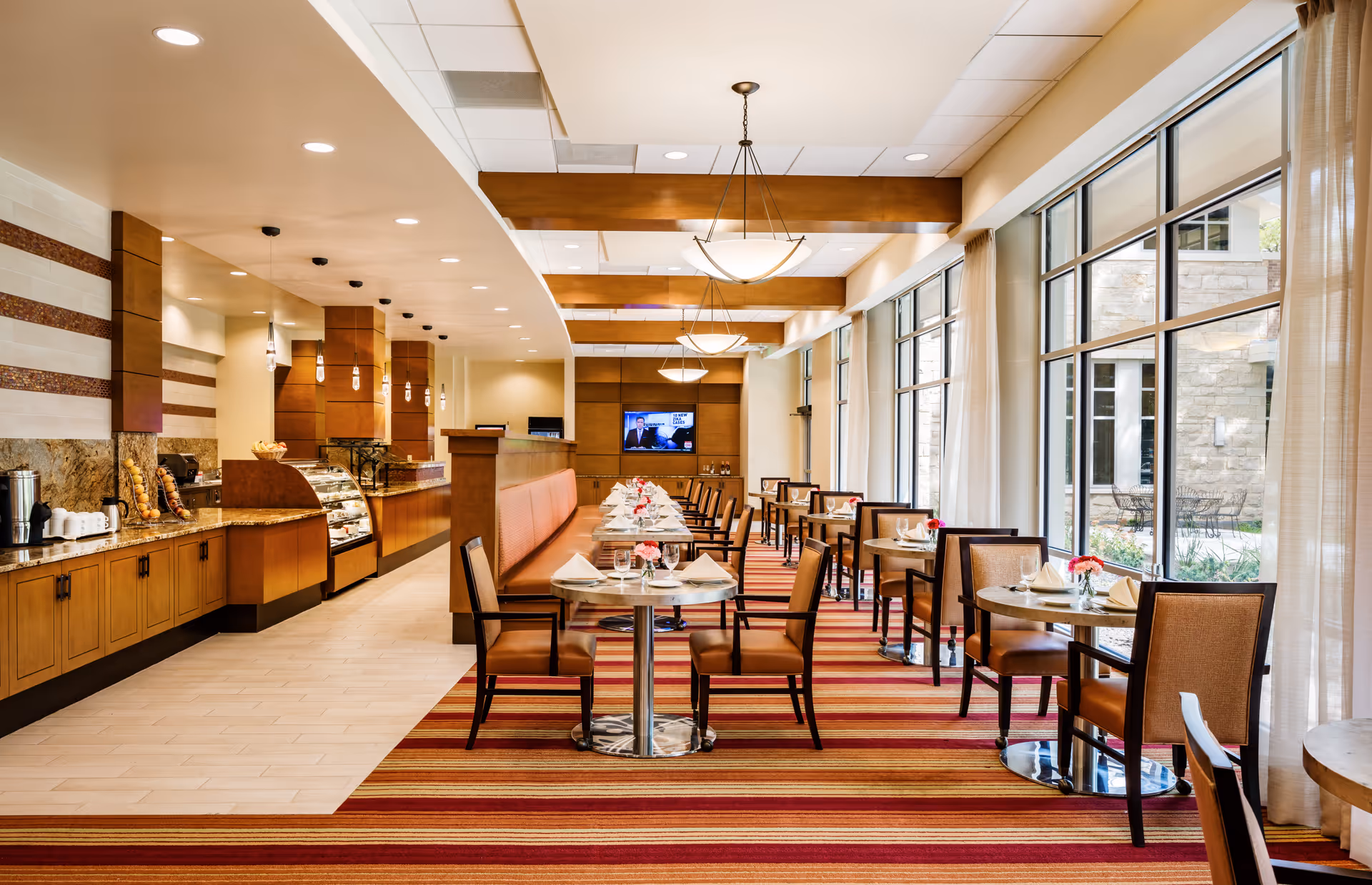 A bright and spacious dining room with large windows on the right side letting in natural light. The room features multiple small round tables with chairs, each set with napkins, glasses, and small flower arrangements. On the left side, there is a long counter with a display case and hanging pendant lights above it. The floor has a striped red and orange carpet, and the ceiling has wooden beams with hanging light fixtures.
