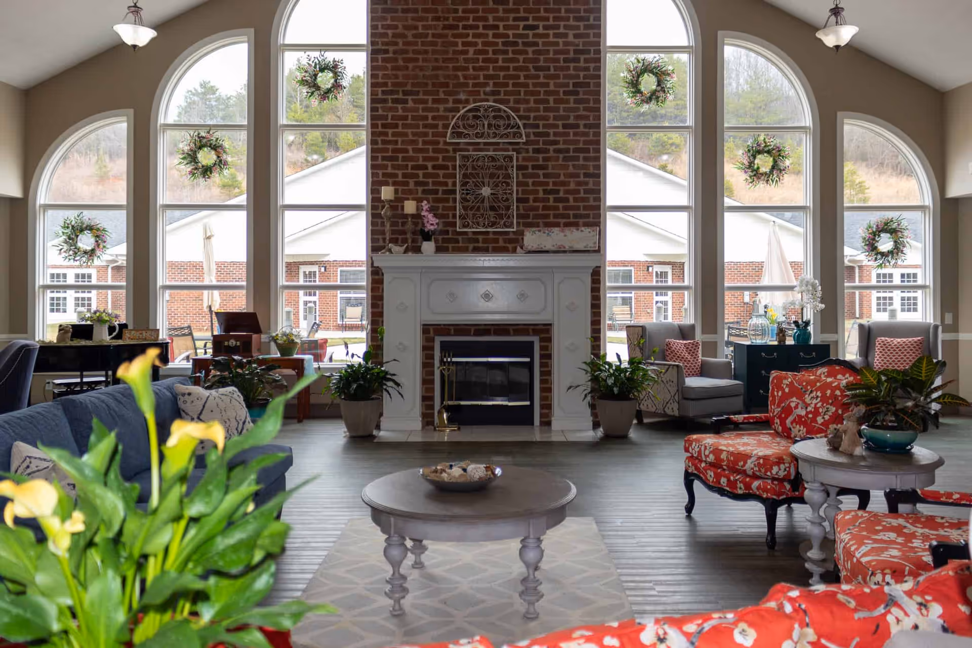 Spacious communal living room with tall arched windows, a central brick fireplace, seating groups, and indoor plants.