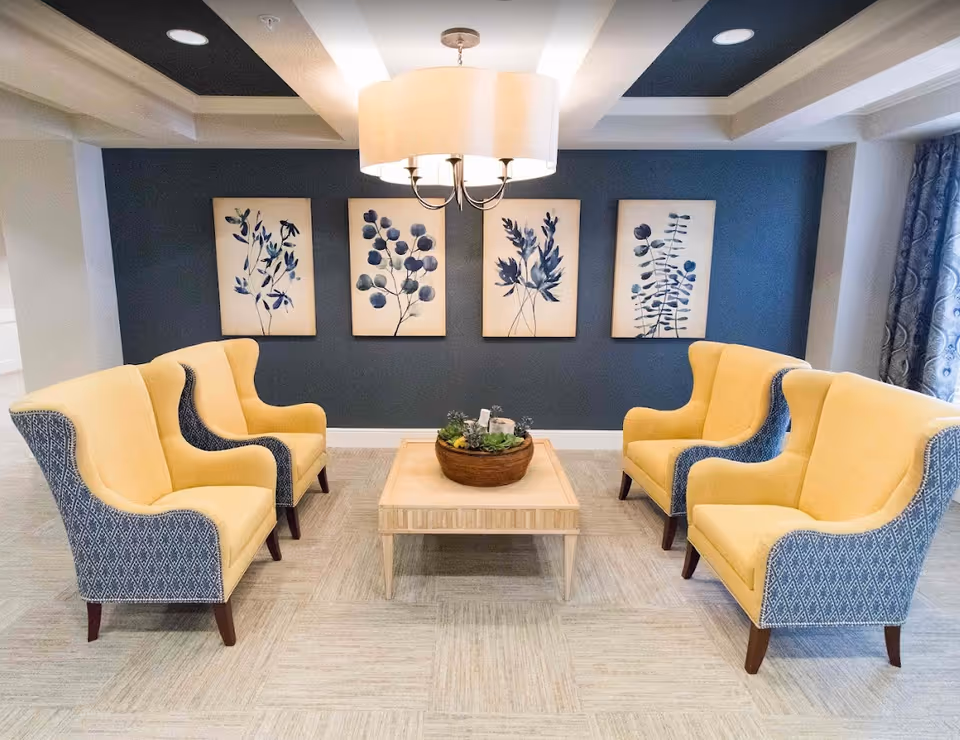 A cozy seating area with four yellow and blue patterned armchairs arranged around a light wooden coffee table with a decorative plant centerpiece. The background features a dark blue wall with four framed botanical prints in shades of blue. A modern ceiling light fixture hangs above the seating area.