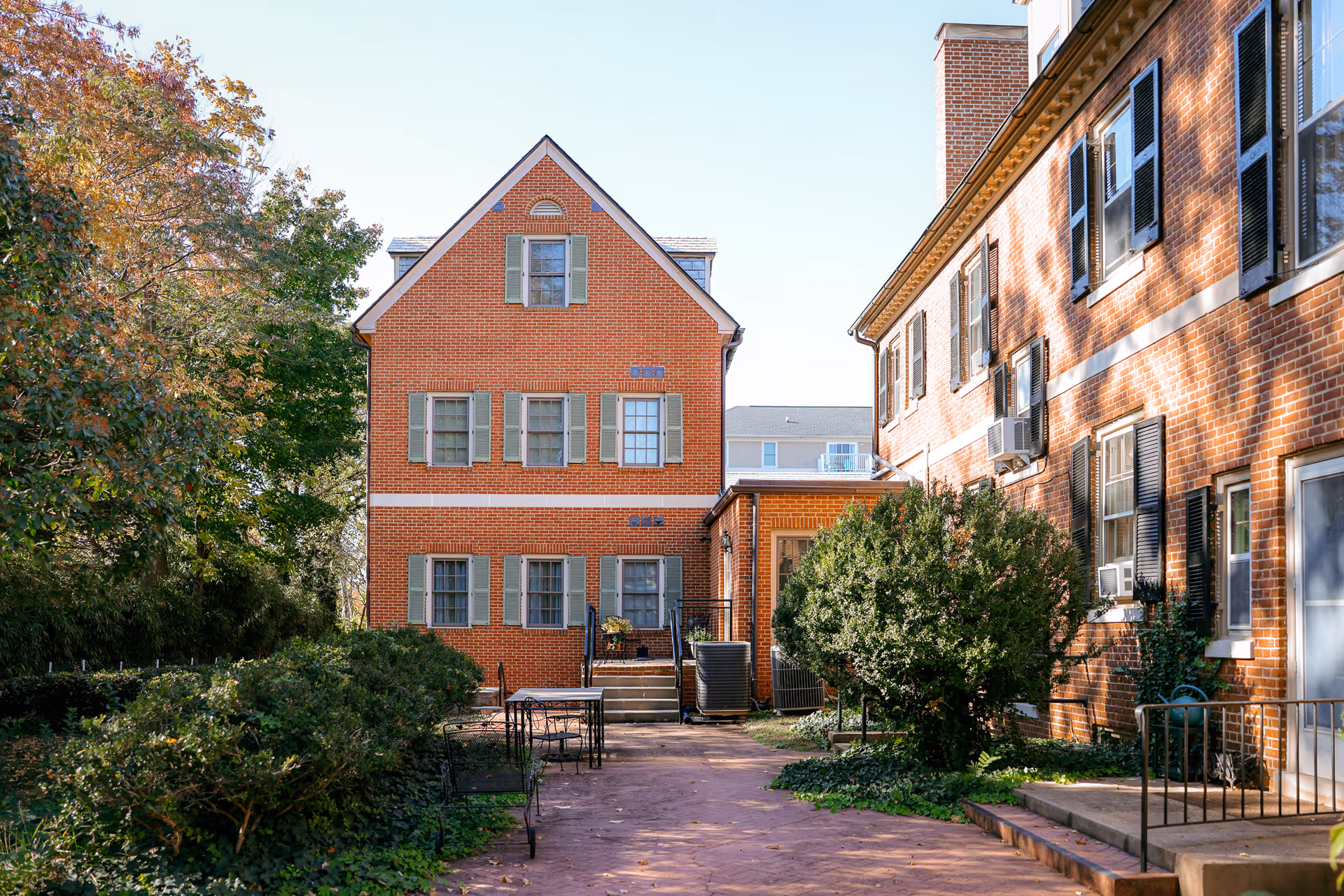 Brick multi-story residential building with a courtyard, shrubs, patio tables, and a paved walkway.