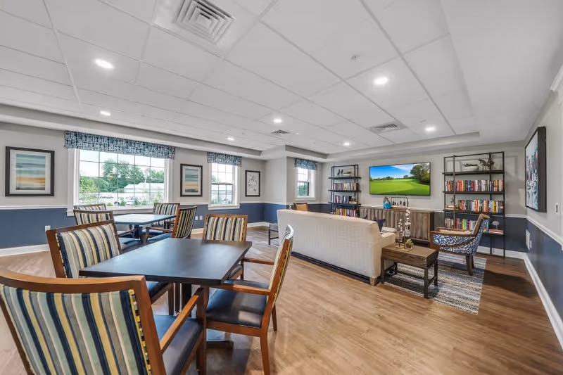 Bright communal lounge with tables and striped chairs, a seating area with a sofa, TV, bookshelves, and large windows.
