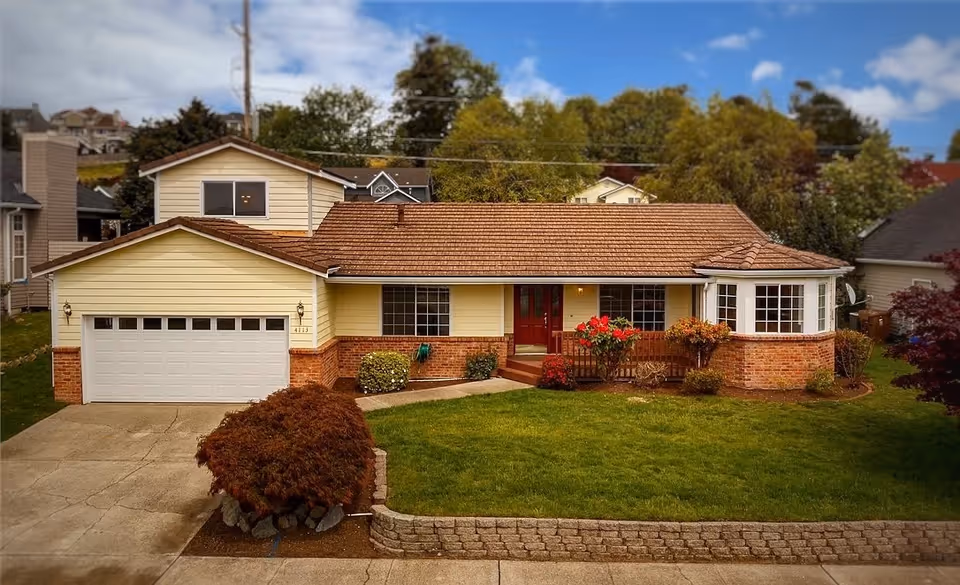 Front exterior view of a single-story house with a two-car garage, a well-maintained lawn, shrubs, and a clear blue sky with some clouds.