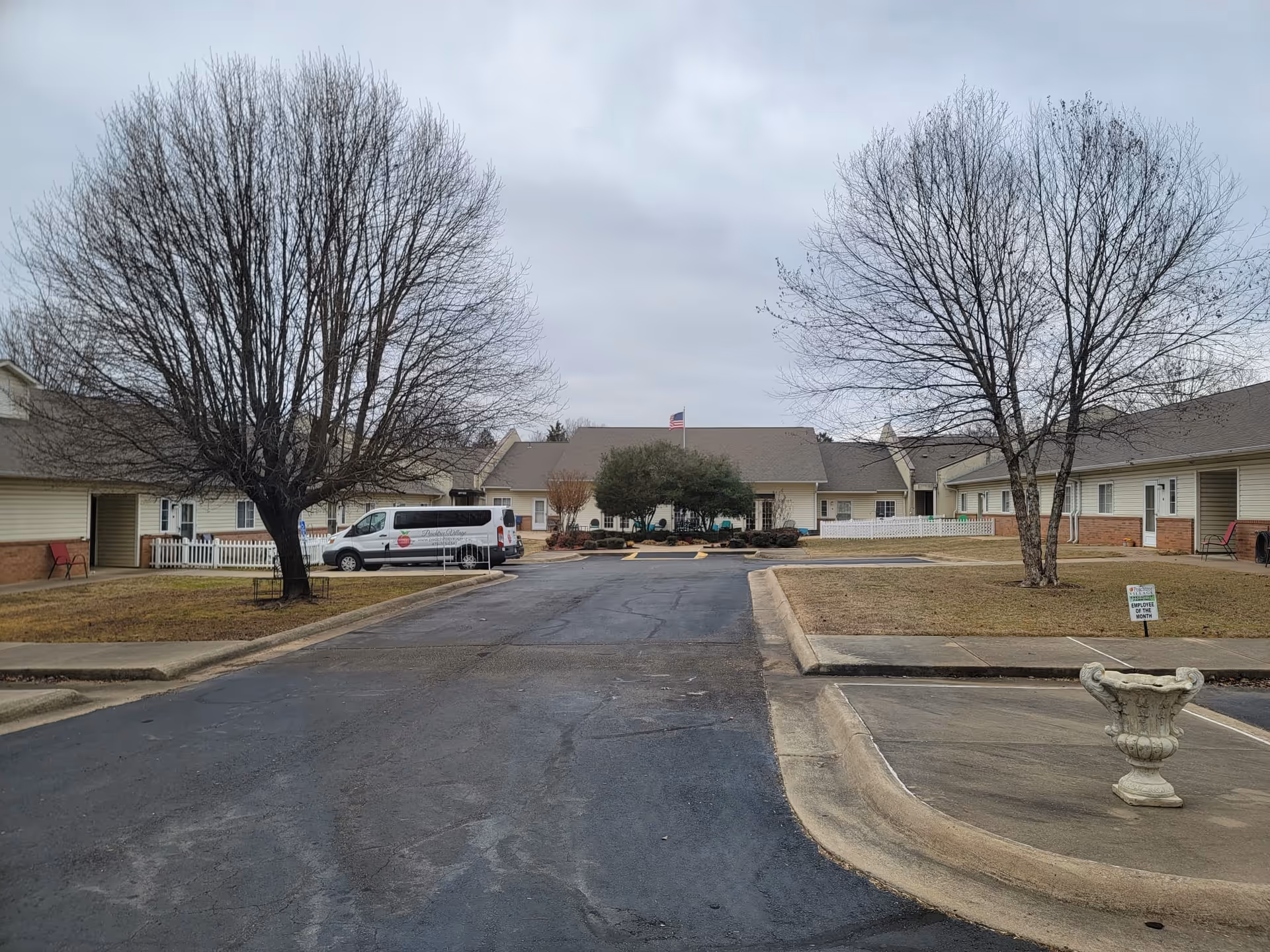 Front exterior of a single-story assisted living facility with a driveway, leafless trees, a parked van, and an American flag above the entrance.