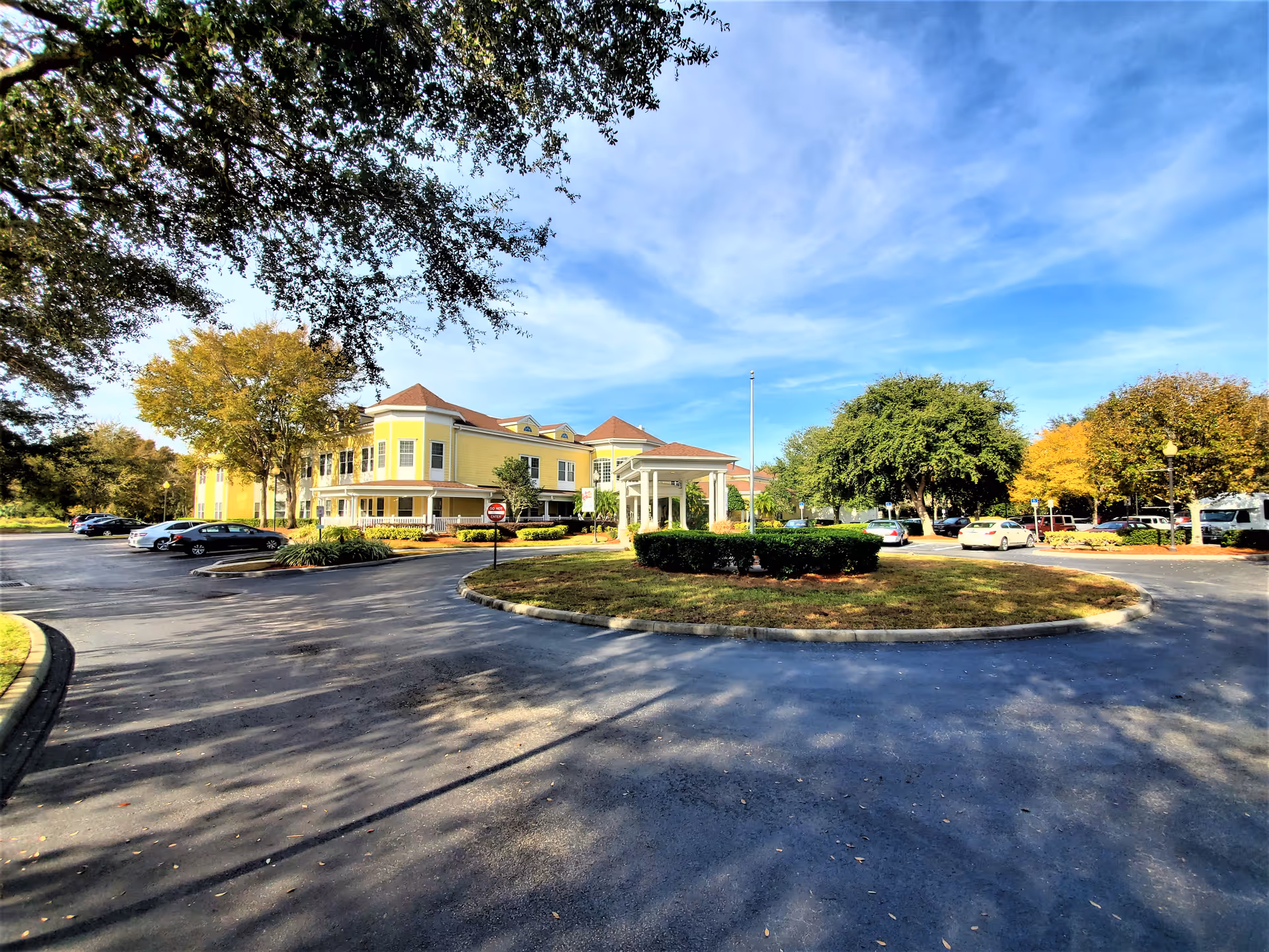 Exterior view of a senior living facility named Madison at Ocoee, showing a yellow two-story building with a covered entrance, surrounded by a circular driveway and parking lot with several cars. Trees with autumn foliage and a bright blue sky with wispy clouds are visible.