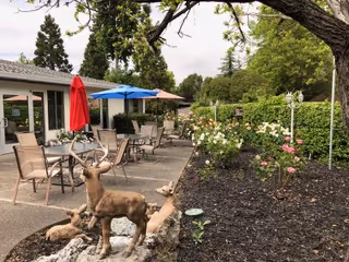 Outdoor patio area with several tables and chairs, some shaded by red and blue umbrellas. A decorative bronze deer statue is positioned near a small rock garden in the foreground. The patio is adjacent to a building with large windows, and there is a garden bed with blooming flowers and greenery along the edge.