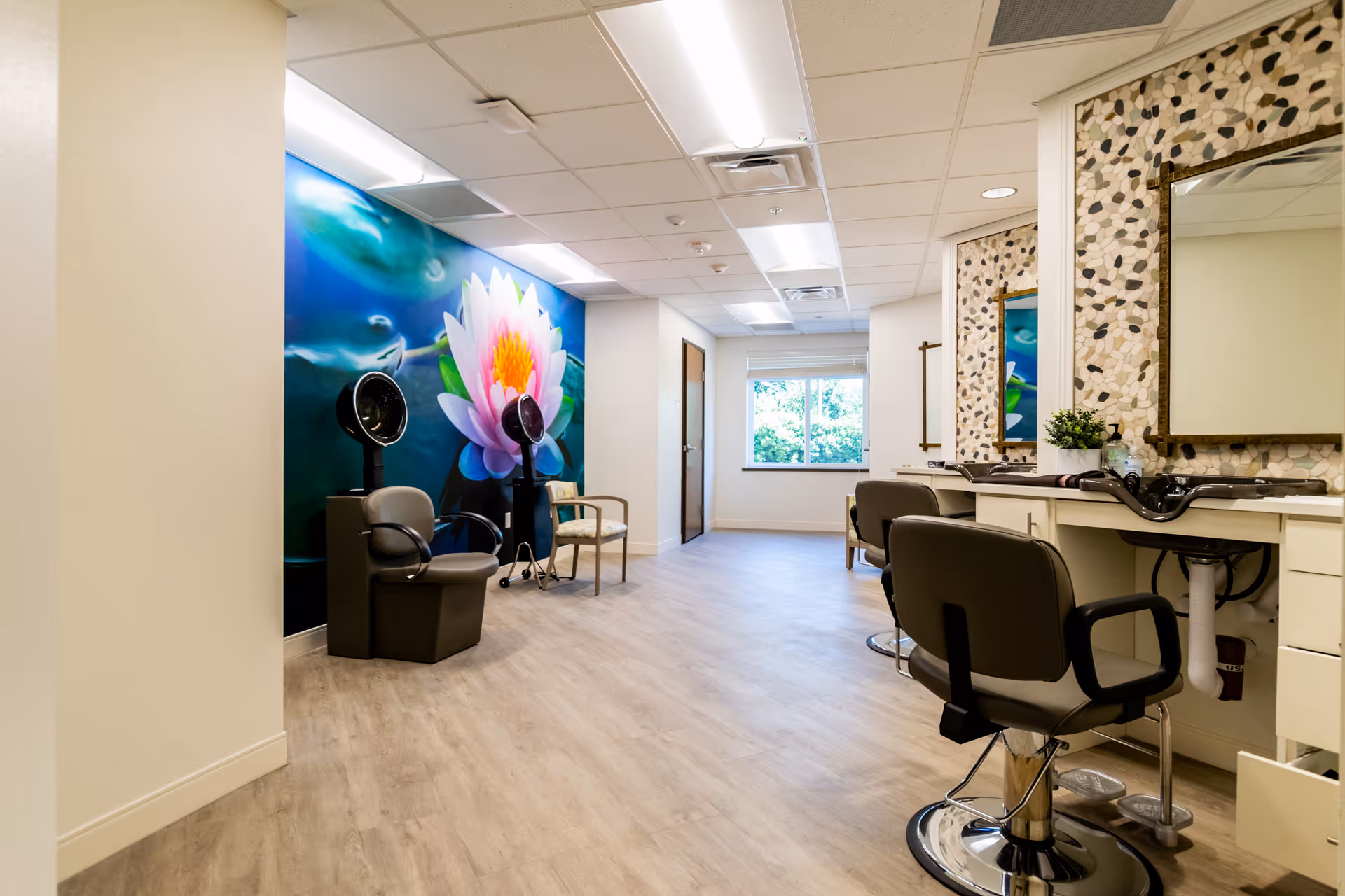 Interior view of a salon area in a senior living facility with hair styling chairs, mirrors, and hair dryers. The room features a large wall mural of a pink and white water lily flower on a blue background and has light wood flooring and bright overhead lighting.