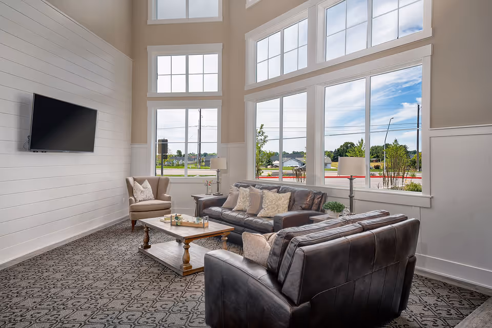A bright and spacious living room area with large windows letting in natural light. The room features two dark leather sofas, a beige armchair, a wooden coffee table with decorative items, and two floor lamps. A flat-screen TV is mounted on a white shiplap wall. Outside the windows, a suburban street and greenery are visible.