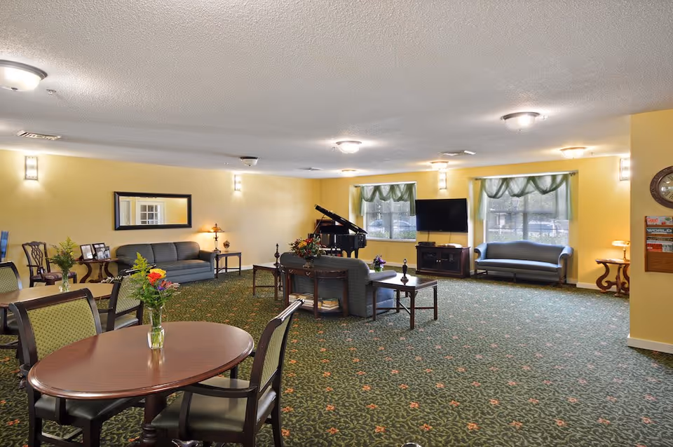 A spacious living room area in a senior living facility with yellow walls and green patterned carpet. The room features multiple seating areas including sofas, armchairs, and tables with chairs. There is a grand piano in the corner, a flat-screen TV on a stand, and windows with green valances letting in natural light. The room is softly lit with ceiling and wall lights, and there are small decorative plants and flowers on the tables.