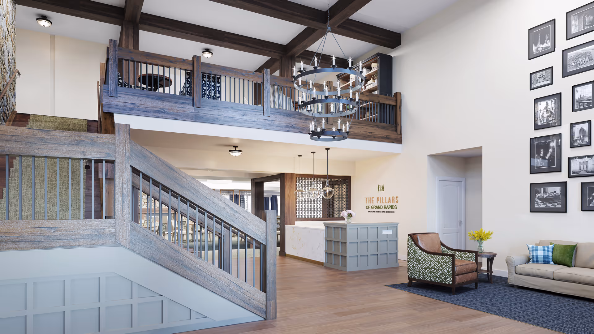 Interior view of The Pillars of Grand Rapids senior living facility featuring a spacious lobby with wooden staircase and railing, a reception desk with pendant lights, a seating area with a sofa and armchair, and a wall decorated with framed black and white photographs.