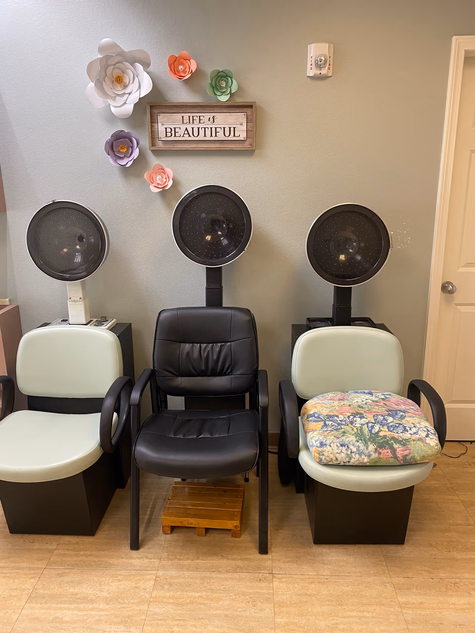 Three salon hair dryer chairs arranged side by side against a light green wall. Above the chairs, there is a wooden sign that reads 'LIFE is BEAUTIFUL' and decorative paper flowers in white, purple, pink, and green. The floor is light wood, and there is a closed door to the right.