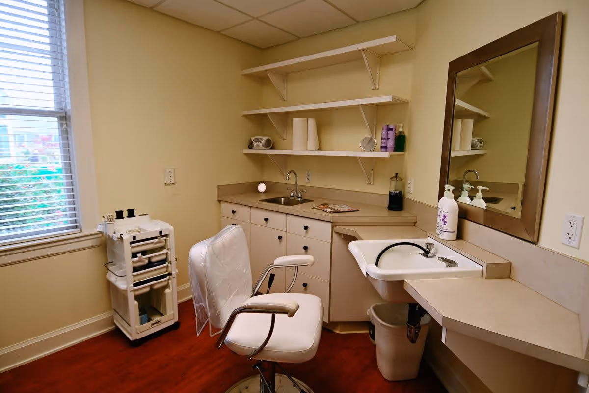 A small room with a white salon chair covered in plastic, a countertop with a small sink, shelves holding paper towels and hair products, a large mirror on the wall, and a window with blinds letting in natural light. There is also a rolling cart with various supplies and a trash bin under the counter.