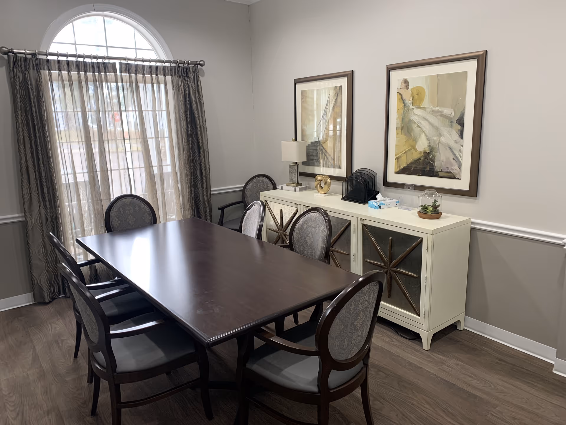 A dining room with a dark wooden rectangular table surrounded by six upholstered chairs. Behind the table is a white sideboard with decorative star designs on the doors, topped with a lamp, tissue box, and small plant. Two framed paintings hang on the wall above the sideboard. A large window with an arched top and sheer curtains allows natural light into the room.