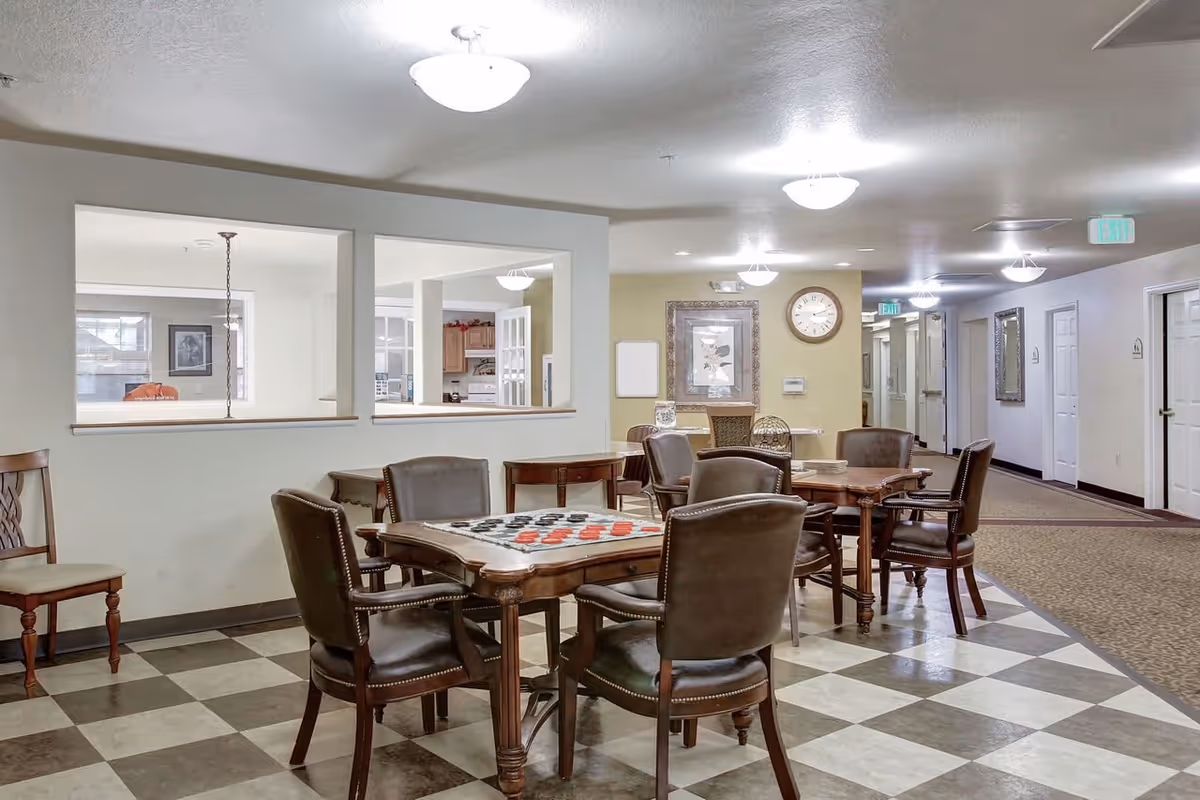 A common area in a senior living facility featuring a checkered floor with several wooden tables and leather chairs arranged for games or socializing. One table has a checkers board set up. The background shows a hallway with doors, framed artwork, a clock on the wall, and windows looking into a kitchen area.