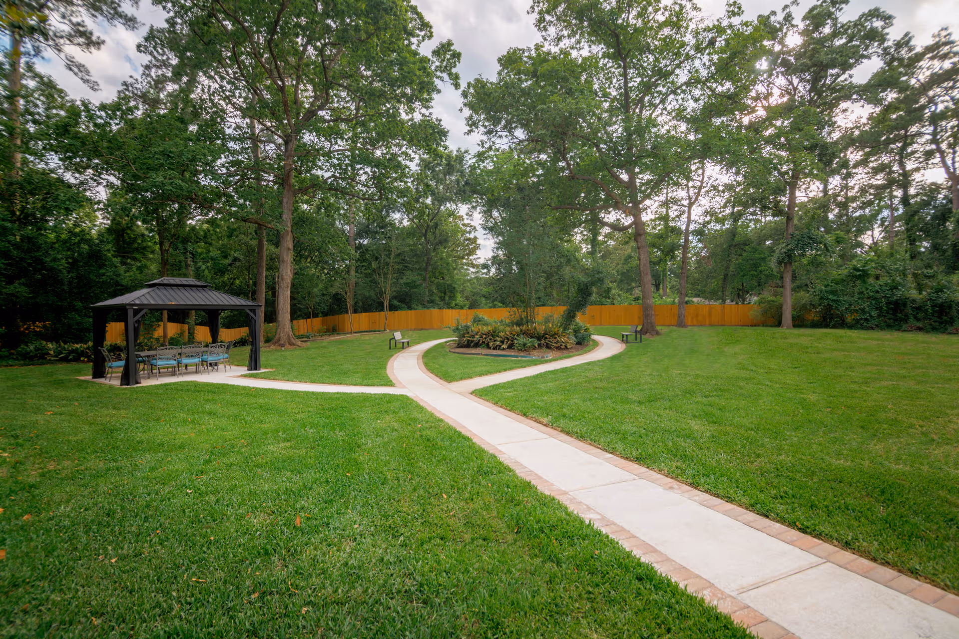 A well-maintained outdoor garden area with a paved walkway splitting into two paths around a central flower bed. There are several large trees, a wooden fence in the background, and a covered seating area with a table and chairs on the left side.