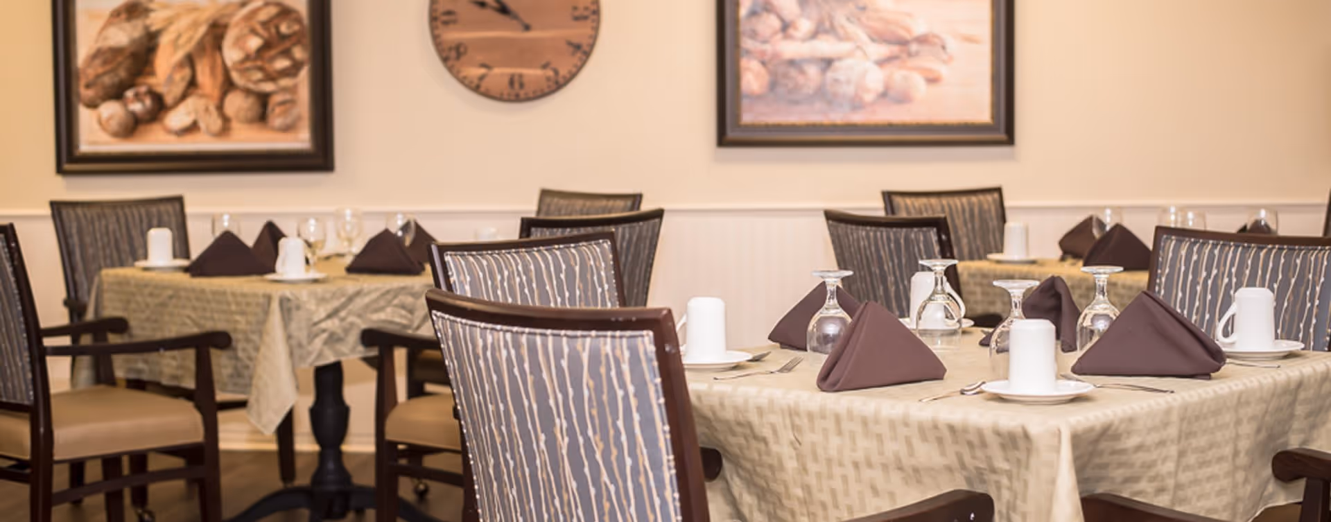 A dining room with tables covered in beige tablecloths, each set with brown folded napkins, upside-down wine glasses, white coffee cups on saucers, and silverware. The chairs have patterned upholstery and wooden frames. The walls are decorated with framed pictures of bread and a clock.