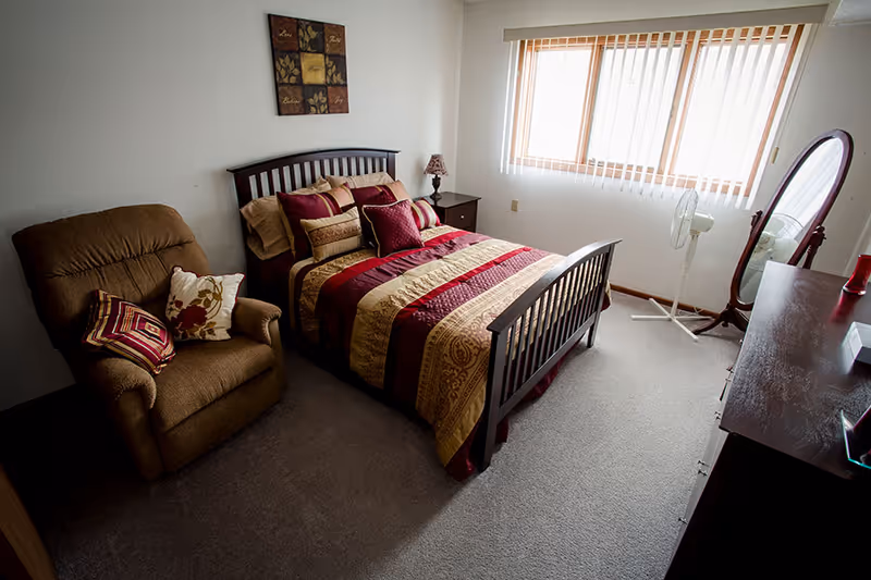 A cozy bedroom with a wooden bed frame featuring a red, gold, and beige patterned comforter and multiple pillows. To the left of the bed is a brown upholstered armchair with two decorative pillows. A nightstand with a lamp is next to the bed. On the right side of the room, there is a standing mirror, a white oscillating fan, and a wooden dresser. A window with vertical blinds allows natural light into the room. A decorative wall hanging is above the bed.
