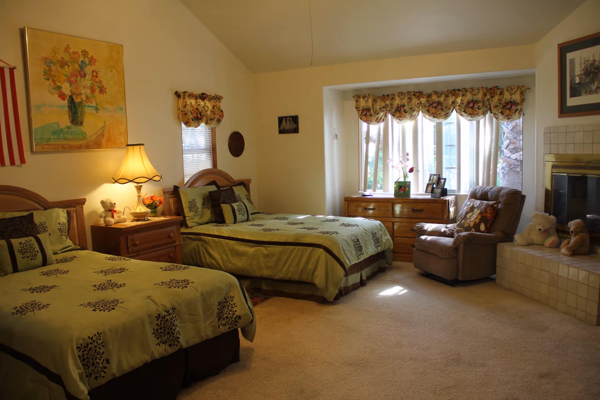 A cozy bedroom with two twin beds covered in matching green and brown bedding. Between the beds is a wooden nightstand with a lamp, a teddy bear, and a small flower arrangement. The room has a large window with floral valances, a wooden dresser beneath the window, and a comfortable armchair next to a tiled fireplace with two stuffed animals on the hearth. The walls are decorated with a painting of flowers and a framed picture.
