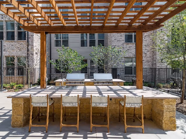 Outdoor seating area with a wooden pergola overhead casting shadows on a stone counter with five wooden chairs. Behind the counter are two built-in grills and some small trees, with a multi-story brick and siding building in the background.