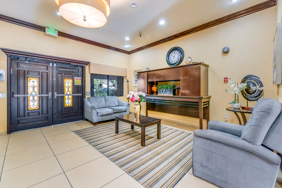 A cozy assisted living facility lobby with a striped area rug, a wooden coffee table with a vase of flowers, two gray upholstered chairs, and a wooden console table with decorative items including a clock and a welcome sign. The entrance features double wooden doors with stained glass panels and an exit sign above.