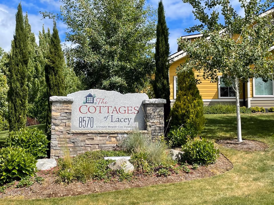 Stone sign for The Cottages of Lacey surrounded by greenery and trees, with a yellow building partially visible in the background under a blue sky with some clouds.