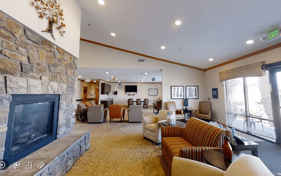 Communal living room with a stone fireplace, armchairs and sofas arranged around tables, and large windows letting in daylight.