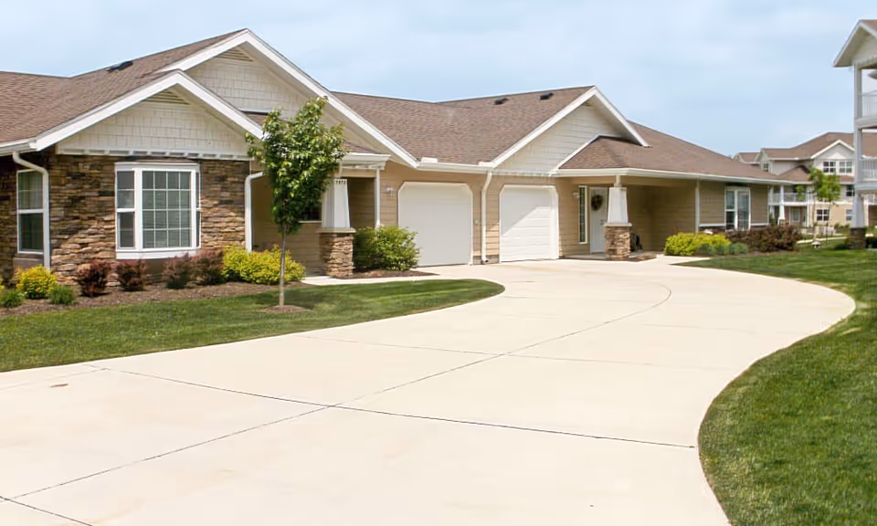 Exterior view of a single-story residential building with a stone and beige siding facade, two white garage doors, a curved concrete driveway, and well-maintained green lawn and shrubs under a partly cloudy sky.