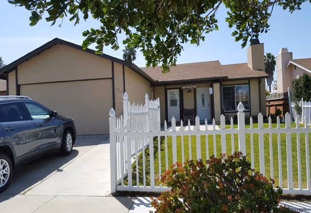 Single-story house with a beige exterior and brown roof, a white picket fence enclosing a green lawn, a driveway with a parked dark-colored SUV, and a tree with green leaves partially shading the front yard under a clear blue sky.
