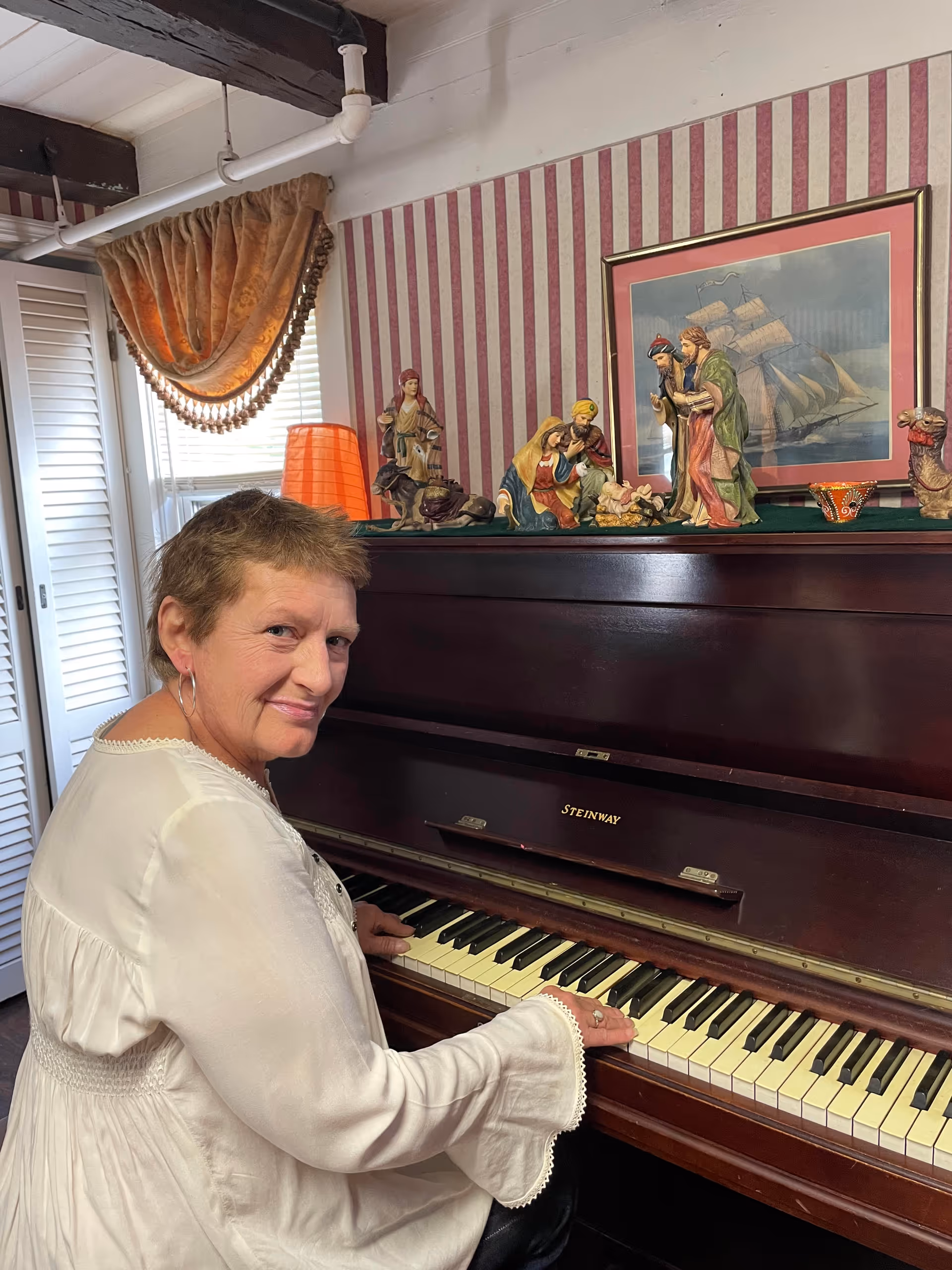 An elderly woman with short hair wearing a white blouse is sitting at a dark wooden Steinway piano, playing the keys and smiling at the camera. Behind the piano, there is a nativity scene with figurines and a framed picture of a sailing ship on a wall with red and white vertical striped wallpaper. A window with white shutters and a decorative curtain is visible to the left.
