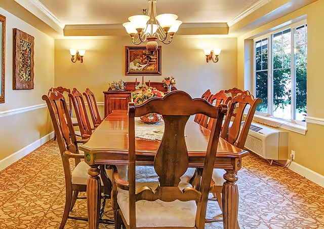 Well-lit formal dining room with a long wooden table, multiple chairs, a chandelier, and a window showing greenery.