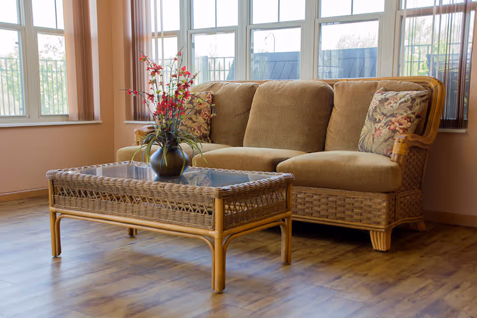 A wicker sofa with floral pillows and a matching glass-top coffee table holding a vase of red flowers in a bright room with large windows.