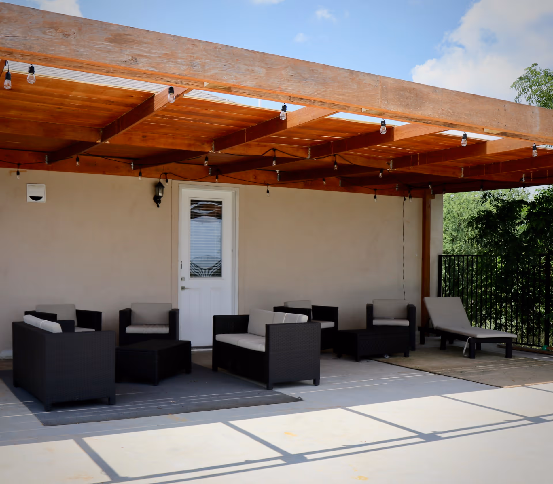 Outdoor patio area with a wooden pergola overhead, featuring several black wicker chairs and sofas with light-colored cushions arranged around a small black coffee table. There is also a lounge chair on the right side. The patio floor is concrete, and there is a white door on the beige wall behind the seating area. Trees and greenery are visible in the background.