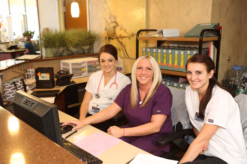 Three female healthcare workers sitting together at a reception desk in a medical or nursing facility. They are smiling and appear friendly. The desk has a computer, paperwork, and office supplies. Behind them are shelves with organized files and office equipment.