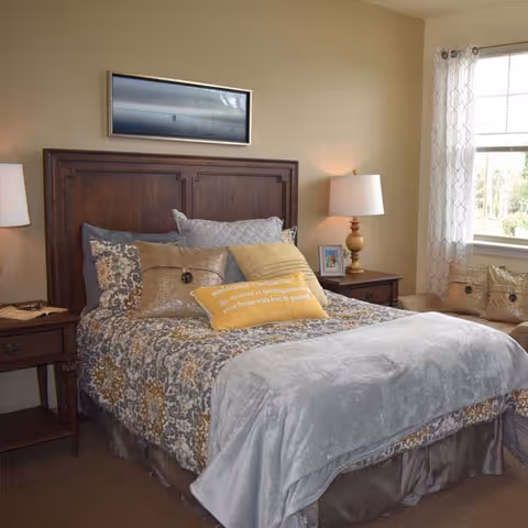A cozy bedroom featuring a wooden headboard bed with patterned bedding in shades of gray, yellow, and white. The bed is adorned with multiple pillows, including a yellow decorative pillow with text. There are two wooden nightstands on either side of the bed, each with a lamp. A window with sheer white curtains allows natural light into the room, and a cushioned bench is placed beneath the window.