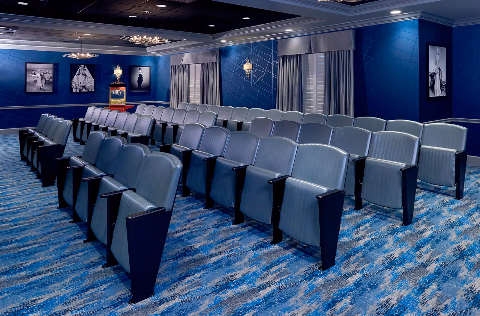 A small theater room with rows of blue cushioned seats arranged in front of a popcorn machine. The walls are painted blue with framed black and white photographs, and there are windows with gray curtains. The carpet has a blue and gray pattern, and the ceiling has recessed lighting and decorative light fixtures.
