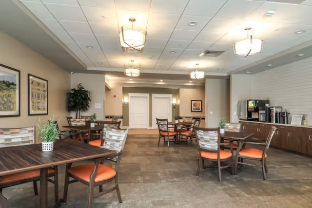 A bright and spacious dining area in a senior living community with several wooden tables and chairs featuring orange cushions. The room has modern ceiling lights, framed artwork on the walls, and a beverage station with coffee dispensers and cups on the right side. There are decorative plants on the tables and in the corner near the wall.