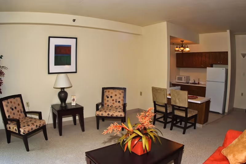 A cozy living area with two patterned armchairs and a small table with a lamp between them. A framed abstract painting hangs on the wall above the table. In the foreground, there is a coffee table with a red vase containing orange flowers. To the right, there is a kitchen area with a refrigerator, microwave, and two chairs at a counter.