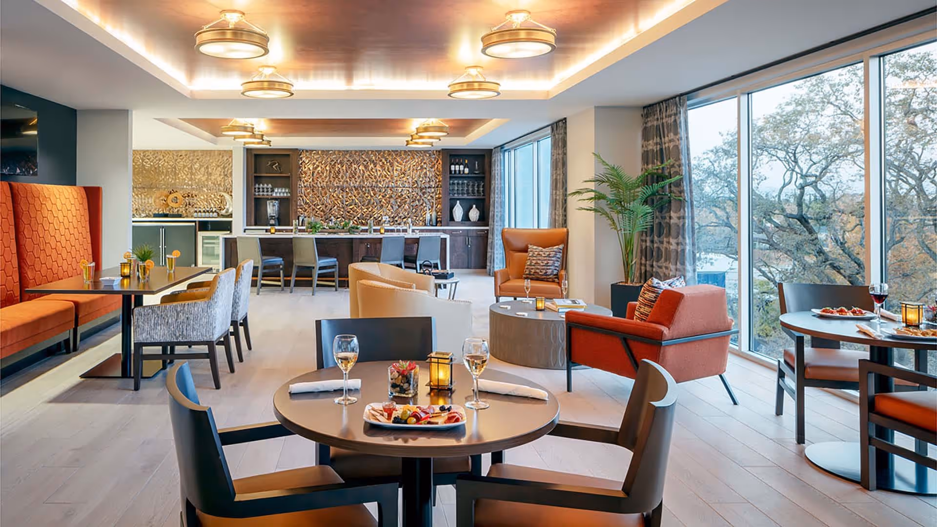 A modern and stylish dining and lounge area with round tables set with wine glasses and plates of food, comfortable chairs, and large windows showing trees outside. The back of the room features a bar with stools and decorative wall art, with warm lighting from ceiling fixtures.