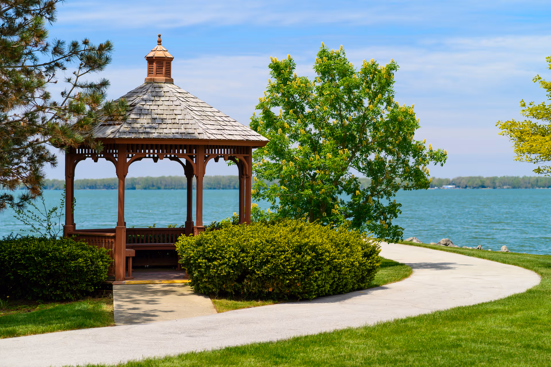 A wooden gazebo with a shingled roof situated near a body of water, surrounded by green bushes, trees, and a curved paved pathway under a blue sky with some clouds.