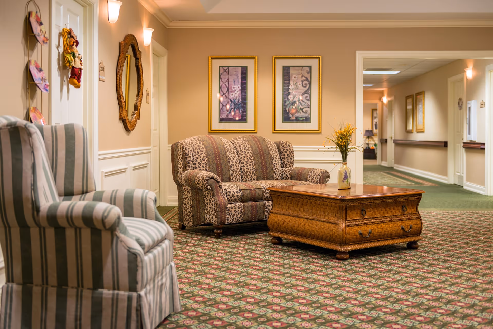 A cozy seating area in a hallway of a senior living facility with a patterned loveseat, a striped armchair, and a wooden coffee table with a vase of flowers. The walls are beige with white trim, decorated with framed floral artwork and a decorative mirror. The carpet has a floral pattern, and the hallway extends into the background with additional doors and wall decorations.