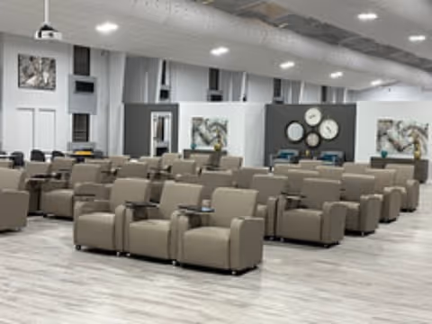 A large, modern common area with multiple rows of beige armchairs arranged neatly on a light wood floor. The room features high ceilings with exposed ductwork and recessed lighting. The walls are decorated with abstract artwork and a set of round clocks displaying different times.