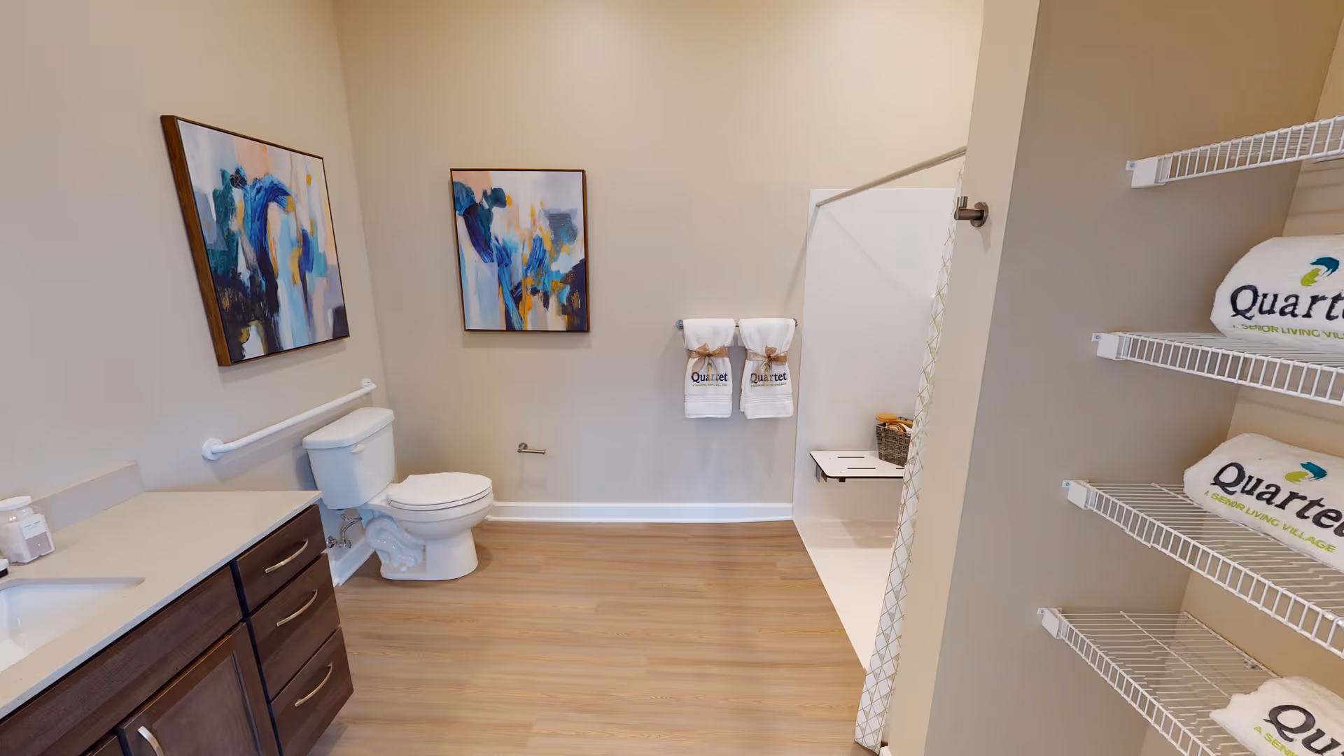 A spacious bathroom with light wood flooring, a white toilet, a shower area with a fold-down seat, and a vanity with a sink and dark wood cabinets. Two abstract paintings hang on the wall above the toilet, and two white towels with the Quartet logo hang on a towel rack. Shelves on the right hold additional white towels with the Quartet logo.