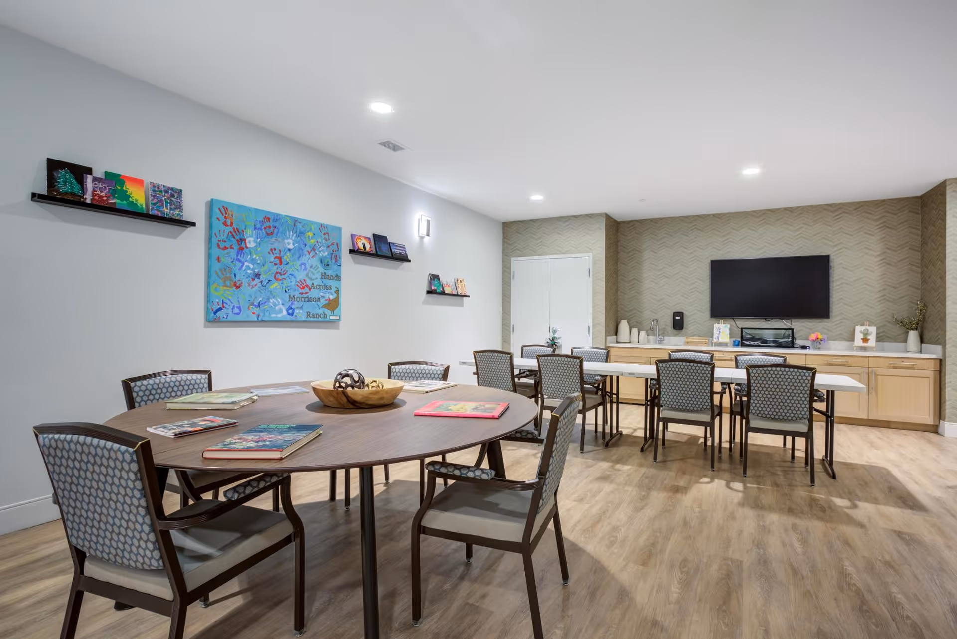 A well-lit common room with two tables surrounded by chairs. The round table in the foreground has books and a decorative bowl on it. The rectangular table in the background has several chairs and is positioned in front of a cabinet with a mounted flat-screen TV above it. The walls are decorated with colorful artwork and shelves holding small items. The floor has a wood-like finish.