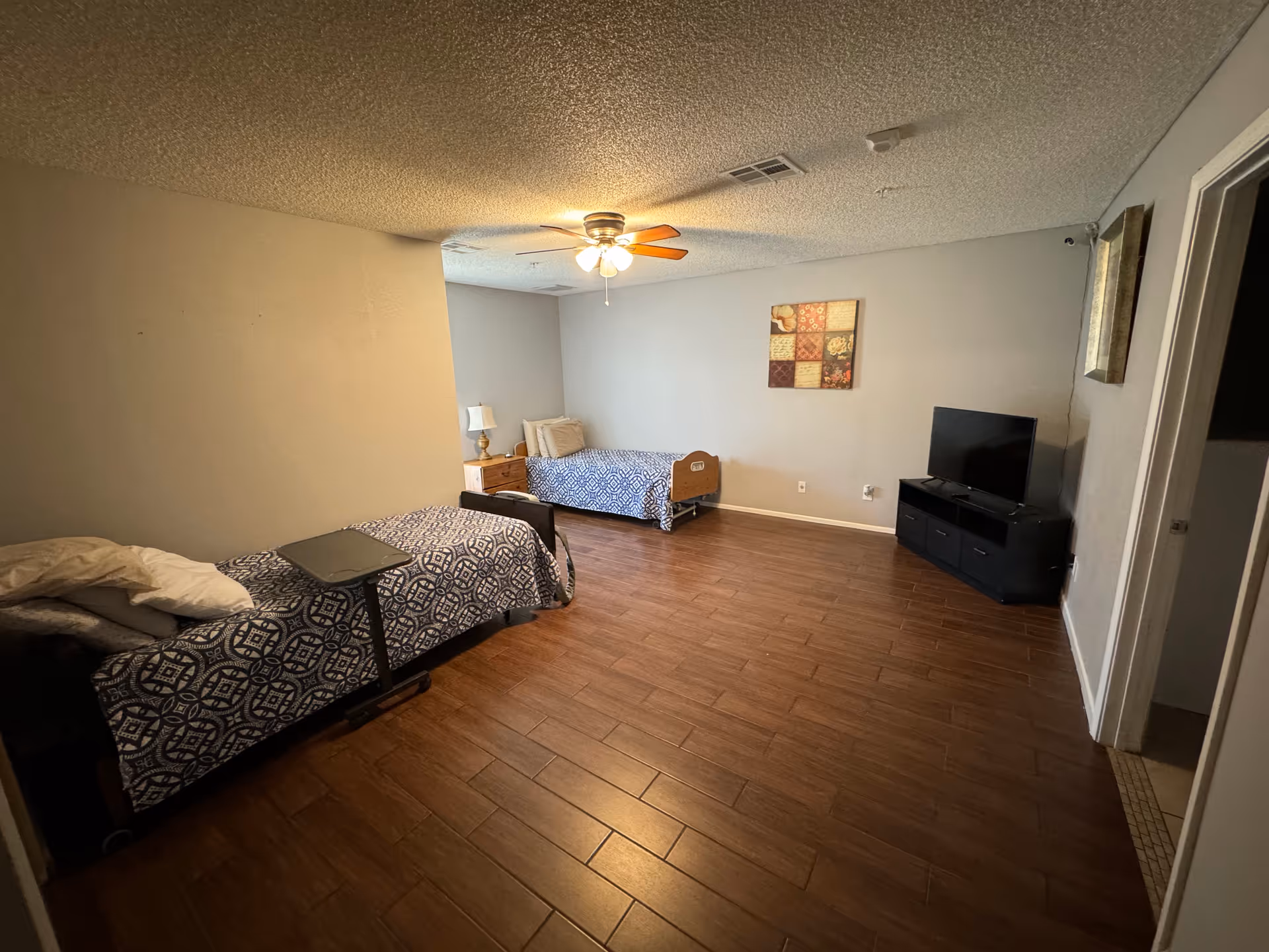 A simple bedroom with two single beds covered in blue patterned bedspreads, a small bedside table with a lamp, a ceiling fan with lights, a flat-screen TV on a black TV stand, and a piece of wall art above one bed. The room has wood-look flooring and light-colored walls.