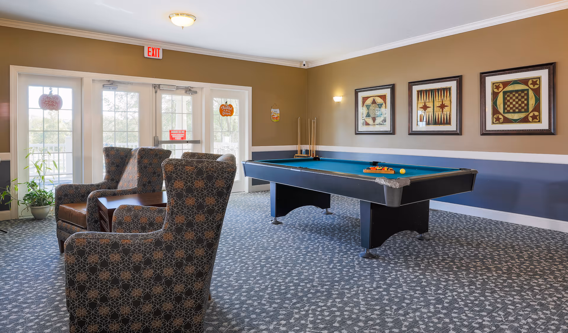 A recreational room with a pool table in the center, surrounded by three patterned armchairs and a small wooden table. The room has carpeted flooring, beige and blue walls with three framed game-themed artworks, and large glass doors letting in natural light. There is a potted plant near the doors and an exit sign above them.